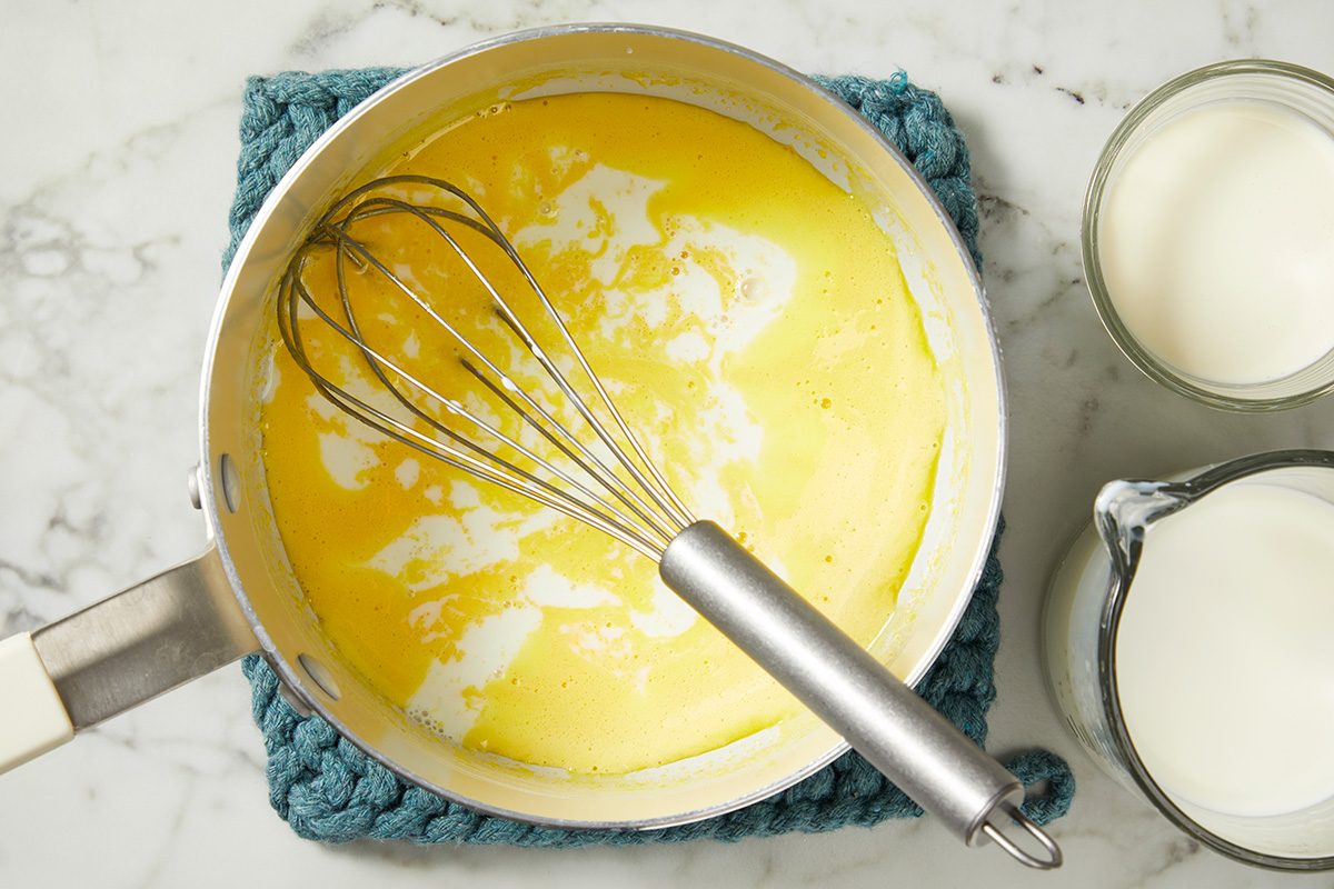 A saucepan filled with melted butter and a whisk on a blue potholder. Two glass containers filled with cream or milk are next to the saucepan on a marble countertop.
