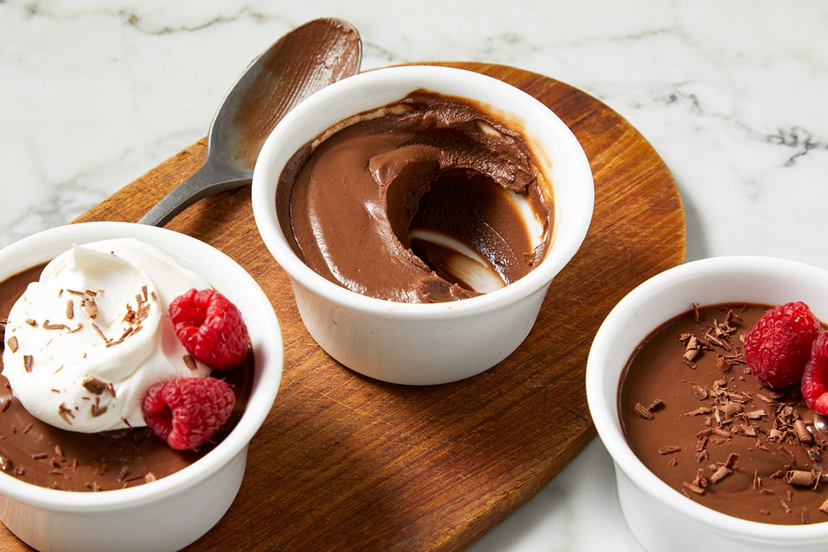 Three ramekins of chocolate mousse sit on a wooden board. One is garnished with whipped cream and raspberries, the second is plain with a spoon, and the third has raspberries and chocolate shavings. The background is a marble surface.