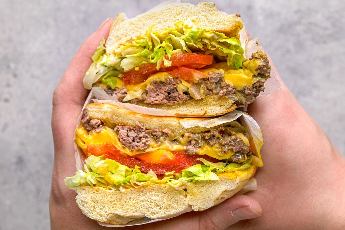 overhead shot of a halved Chopped Cheese sandwich being held in two hands, the sandwich is cut vertically and showcases layers of chopped beef, melted orange cheese, slices of red tomato, and shredded lettuce, all nestled within a soft, white bun, the sandwich is wrapped in white parchment paper at the bottom, and the hands are positioned against a plain, light grey background