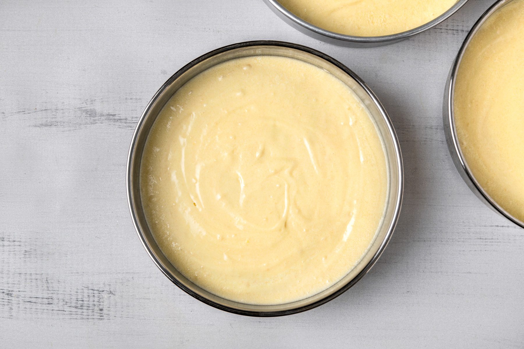 overhead shot of several round baking pans filled with a smooth, pale yellow batter; the pans are metallic and sit against a light grey surface;
