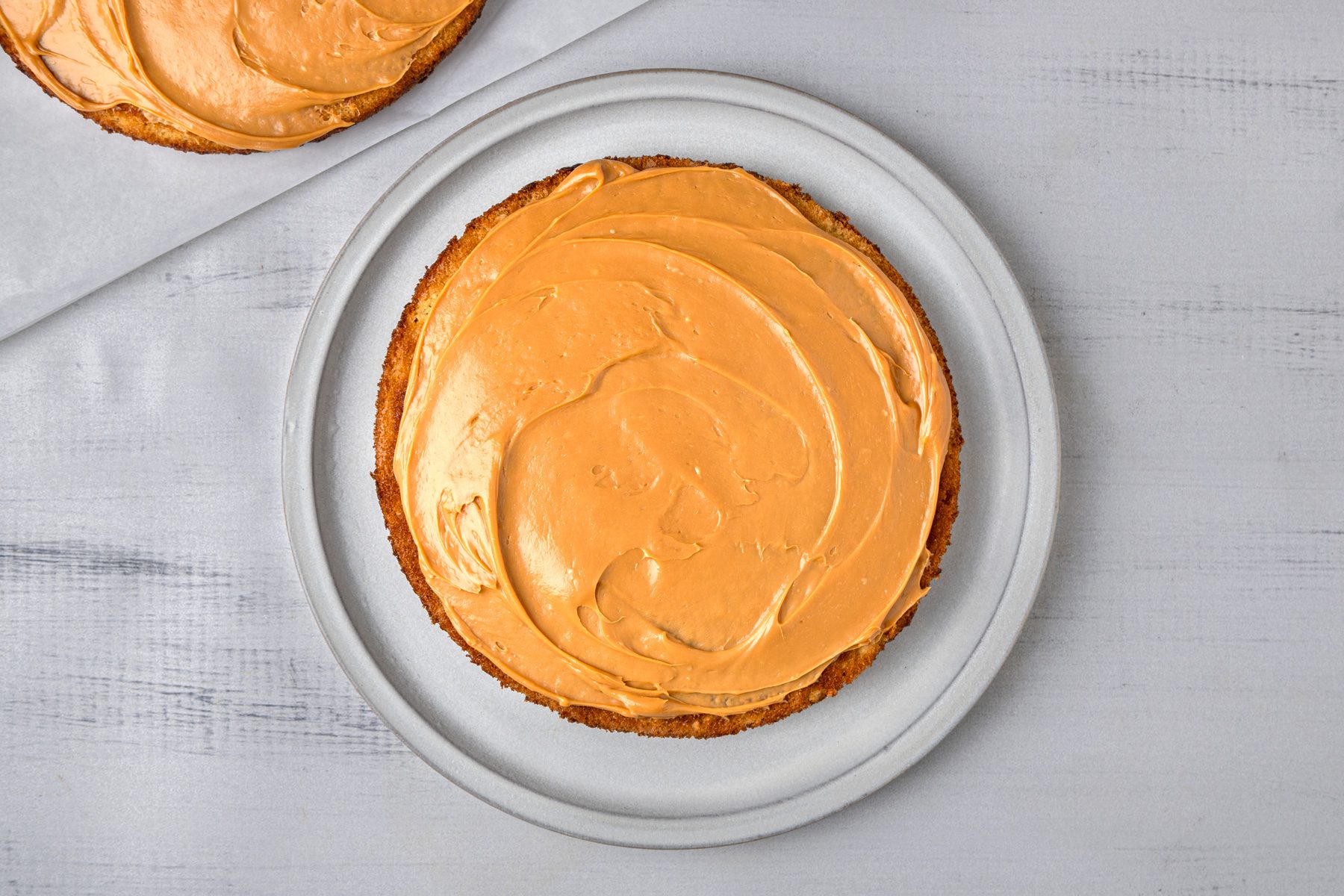overhead shot of a round cake that is displayed on a light grey plate; the cake has a smooth, caramel colored frosting spread evenly across its top, beside the cake, there is another similar cake on a piece of parchment paper that is slightly out of focus