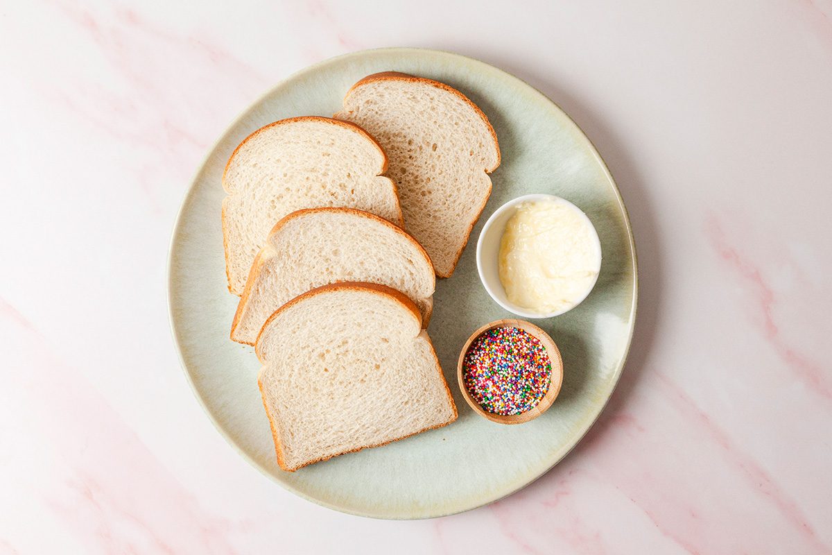 A plate with four slices of white bread, a small bowl of colorful sprinkles, and a small bowl of butter on a light pink marble surface.