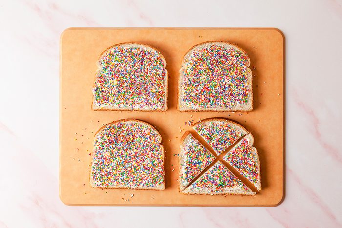 Four slices of bread covered with colorful sprinkles are placed on a wooden cutting board. Three slices are whole, while one is cut into two triangles. The background is a light, marbled surface.