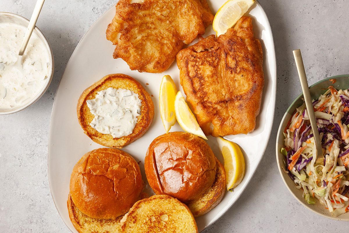 A plate with two pieces of golden-brown fried fish, lemon wedges, and toasted buns. A small bowl of creamy tartar sauce with a spoon is to the left, and a bowl of coleslaw with a spoon is on the right.