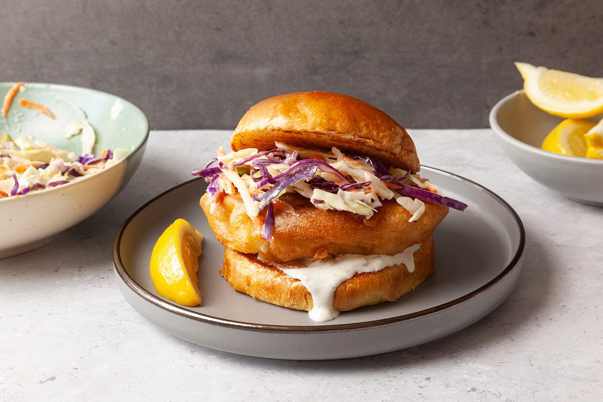 A fish sandwich with coleslaw on a plate, accompanied by a lemon wedge. Two bowls in the background contain more coleslaw and lemon slices. The setting is on a light countertop with a gray background.