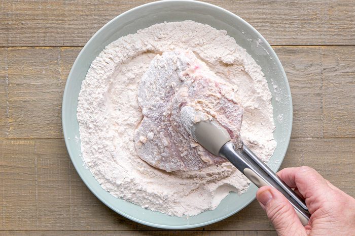 overhead shot of a hand holding a pair of tongs while coating a piece of meat in a flour mixture; the meat appears to be partially submerged in the flour, which is placed in a shallow, round dish; the background consists of wooden surfaces