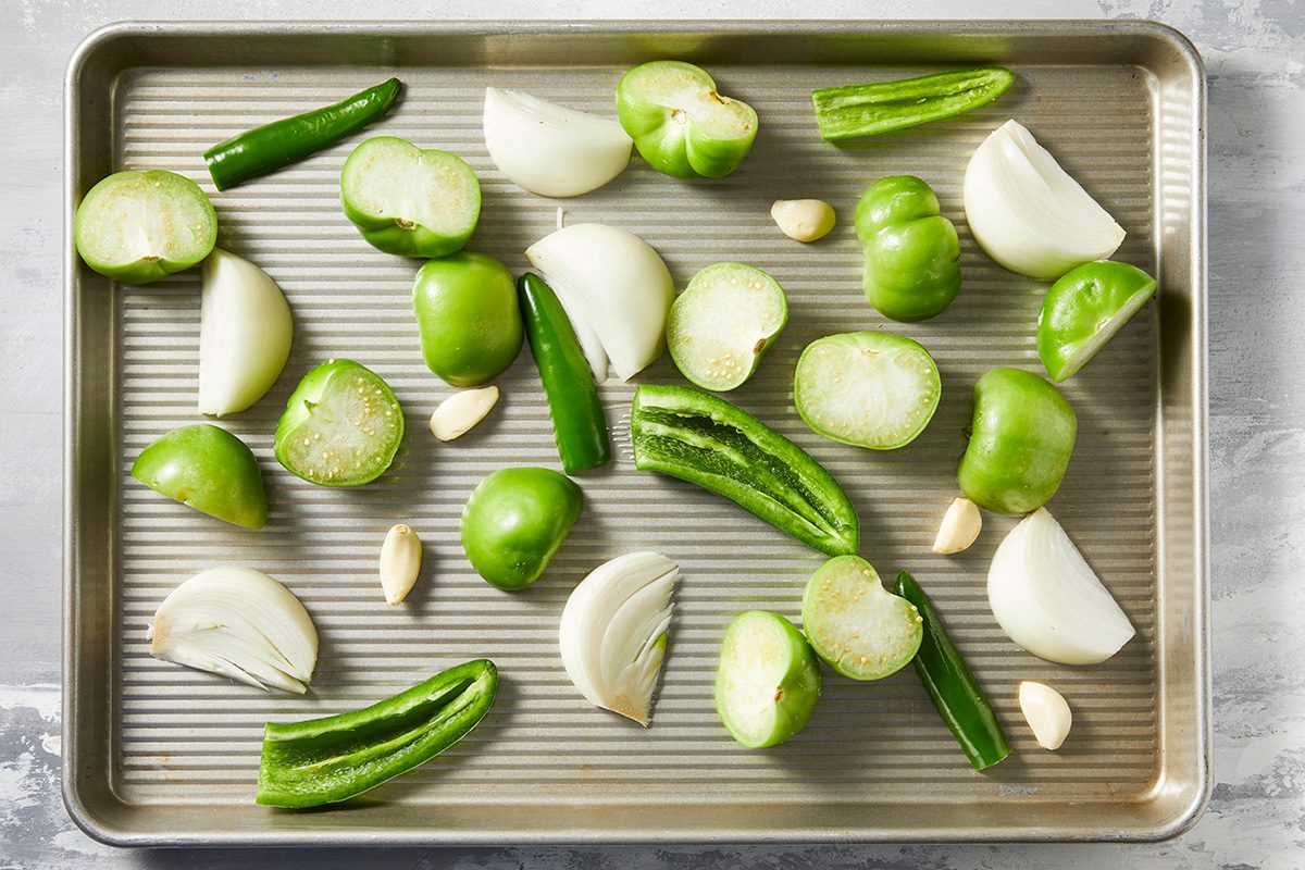 Overhead shot of place tomatillos, chiles, onion, jalapeno and garlic on a rimmed baking sheet; Drizzle with oil; sprinkle with salt; cumin and pepper.; Toss to thoroughly coat