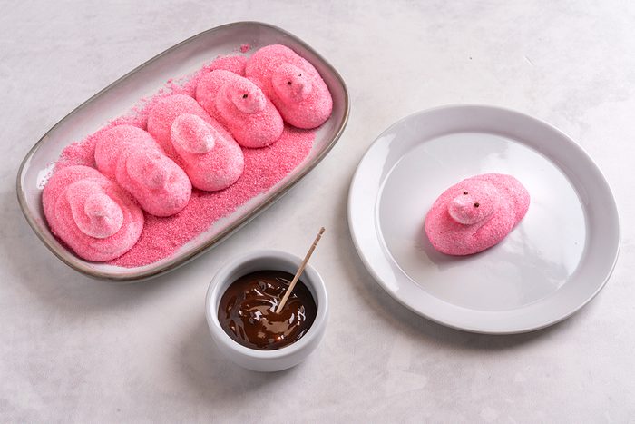 A dish with four pink marshmallow chicks coated in sugar is next to a white plate with one marshmallow chick. A small white bowl filled with chocolate sauce and a toothpick is positioned in front of the plate.