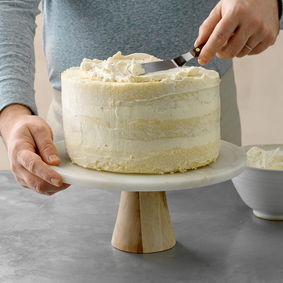 A person is frosting a tall, round cake with white icing on a wooden cake stand. They are using an offset spatula to spread the icing smoothly. A bowl of extra frosting is visible nearby on a gray countertop.