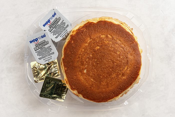 A top view of a packaged meal featuring two golden-brown pancakes, accompanied by packets of syrup and butter on the side, placed on a clear plastic tray.