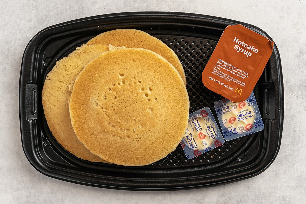 A black tray with three golden-brown pancakes, a container of hotcake syrup, and two butter packets on a light surface.