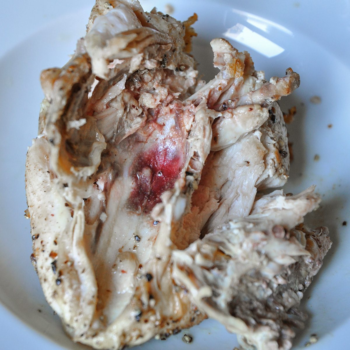 A partially cooked piece of chicken breast on a white plate, showing a raw, pink and red center. It is seasoned with black pepper and herbs.