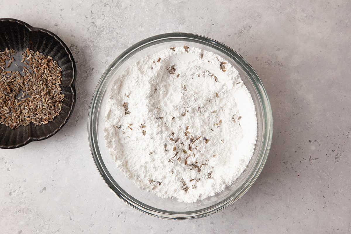 A glass bowl filled with flour and scattered seeds. To the left, a small black dish holds additional seeds. The background is a light gray surface.