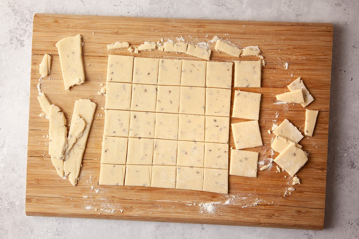 A wooden cutting board with neatly arranged square pieces of dough, dusted with flour. The dough squares are surrounded by trimmed edges and scattered small pieces, all set on a light stone counter.