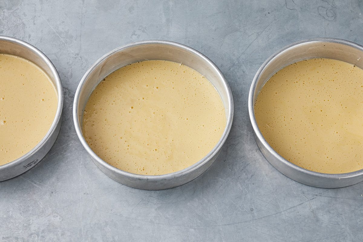 Three round cake pans filled with smooth, light yellow batter are placed side by side on a gray surface.