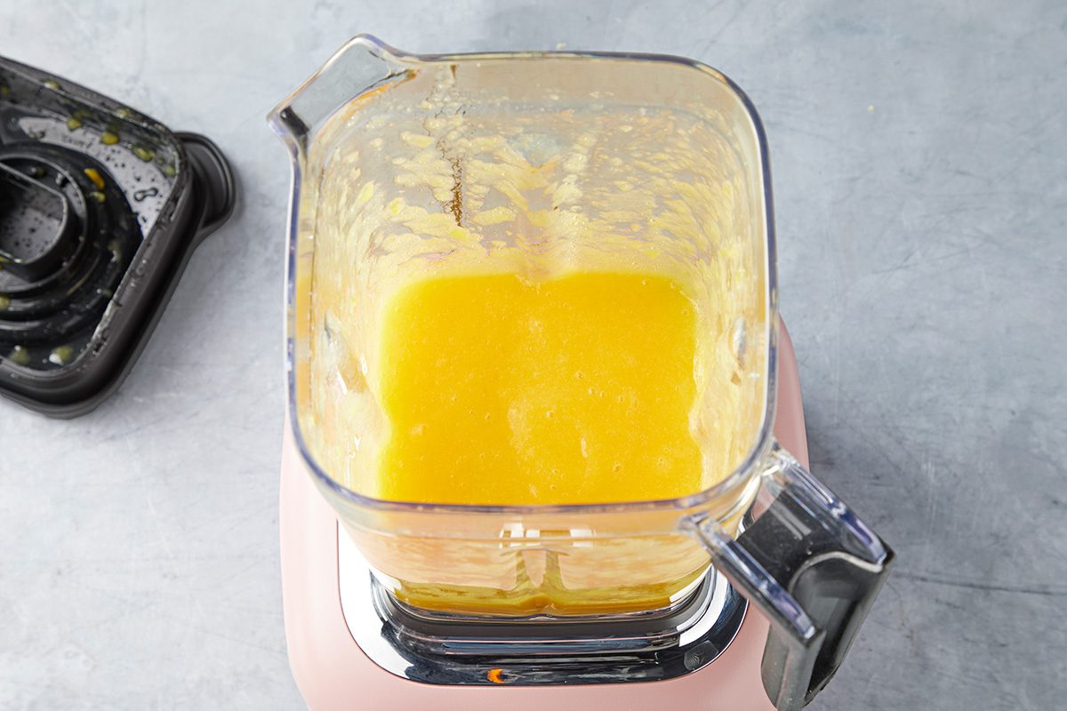 Top view of a blender with bright yellow liquid inside, likely a smoothie or juice, on a gray countertop. The blender lid is placed nearby, slightly stained with remnants of the blend.