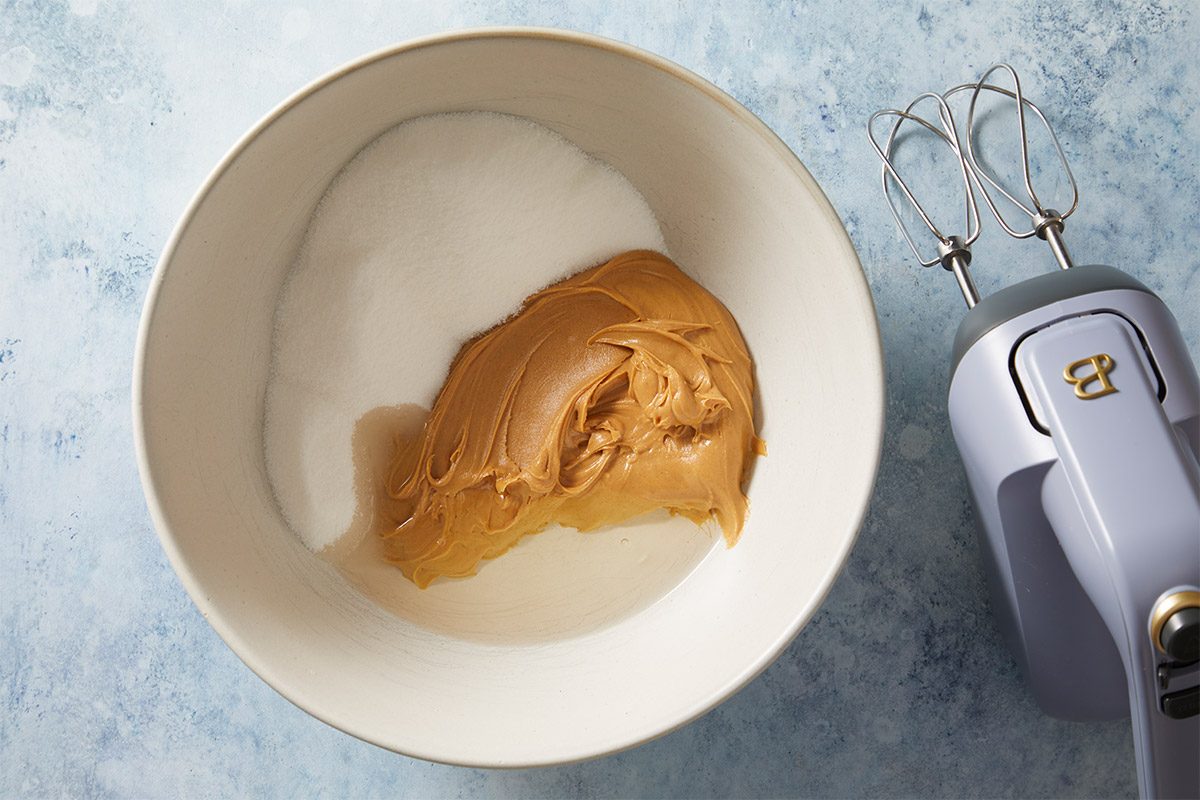 overhead shot of a mixing bowl containing a blob of creamy peanut butter placed next to a mound of white sugar; the bowl is positioned against a light blue countertop, to the right, a handheld electric mixer rests