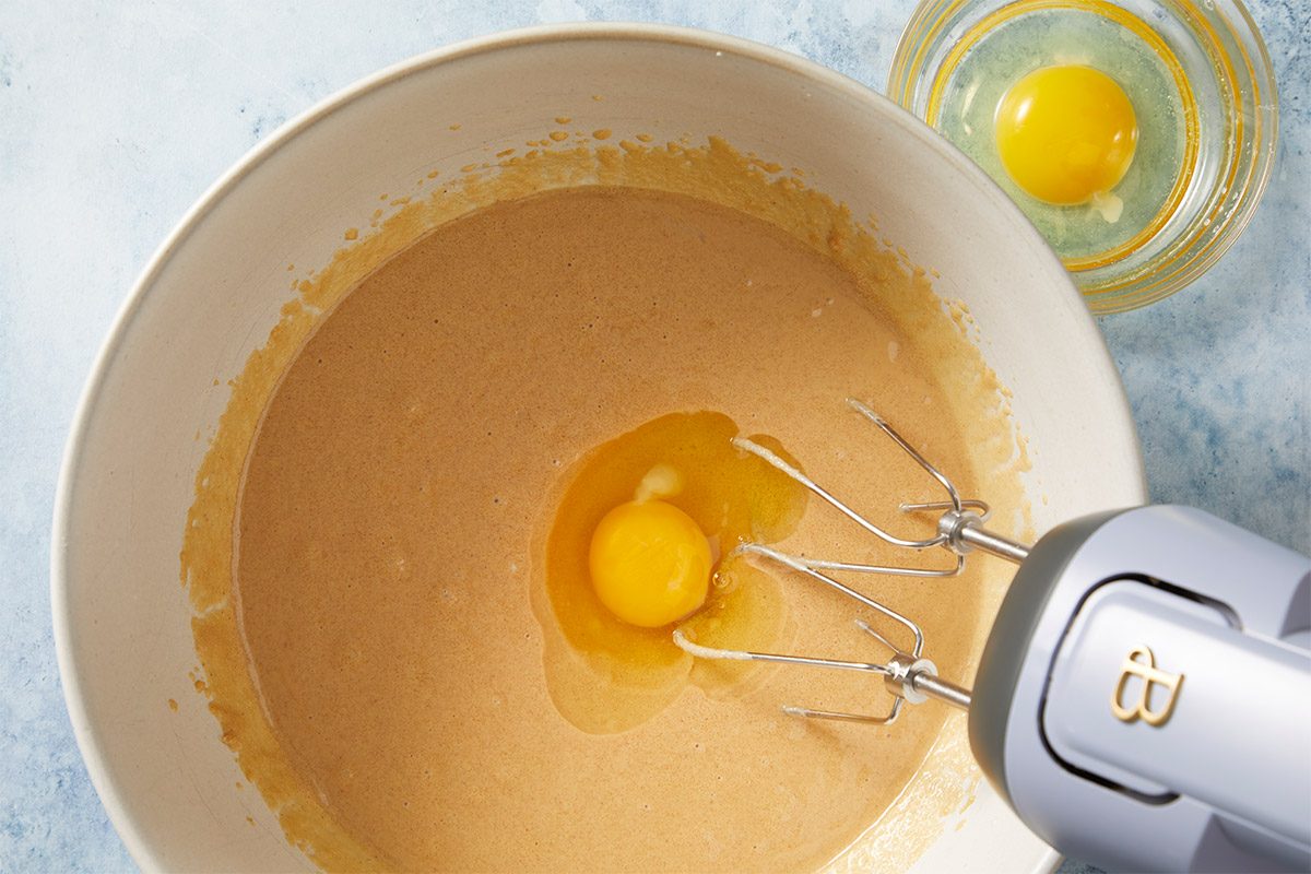 overhead shot of a large mixing bowl filled with a batter; at the center of the batter, there is a raw egg with its yolk visible, beside the bowl, a handheld mixer is positioned, its beaters resting in the batter; a smaller bowl containing another whole egg sits in the background