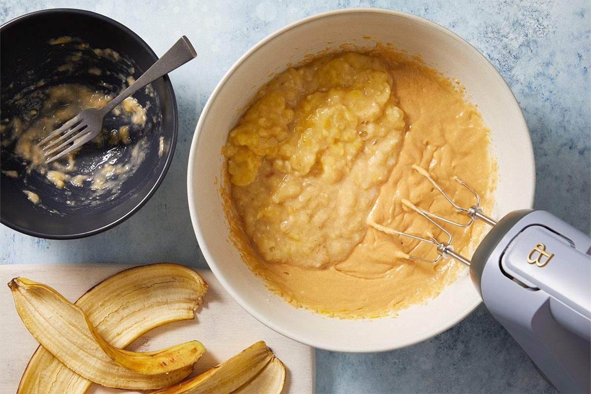 overhead shot of a light colored bowl filled with a thick, creamy mixture; A handheld mixer rests next to the bowl, with its beaters slightly submerged in the mixture; There is a small black bowl nearby containing remnants of mashed banana, and a fork is placed inside it; several banana peels are laid out on a wooden cutting board positioned at the bottom of the image, The background features a soft, light blue color