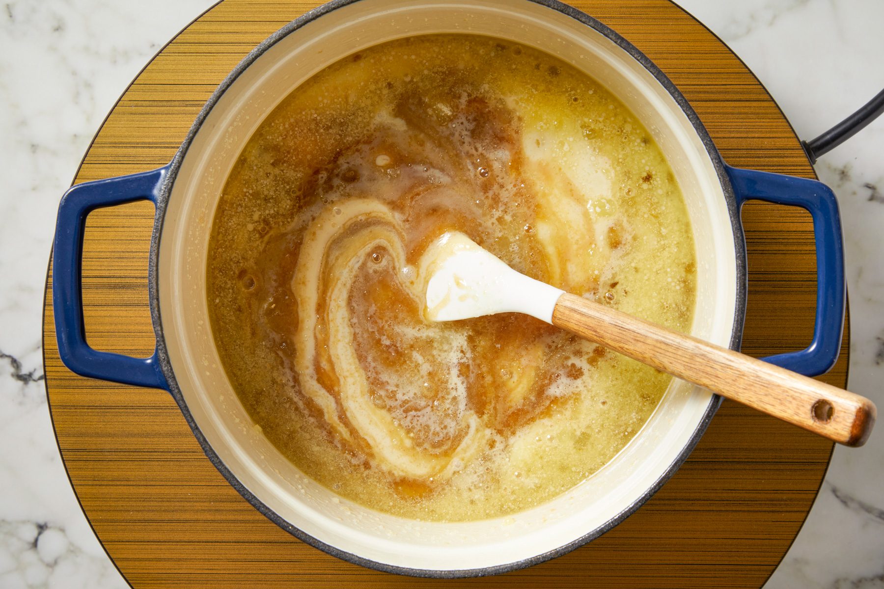 Overhead shot of a large heavy saucepan over medium heat melt cubed butter; Stir in sugars; cream; milk; corn syrup and salt; cook and stir until mixture comes to a boil; spatula; marble surface;