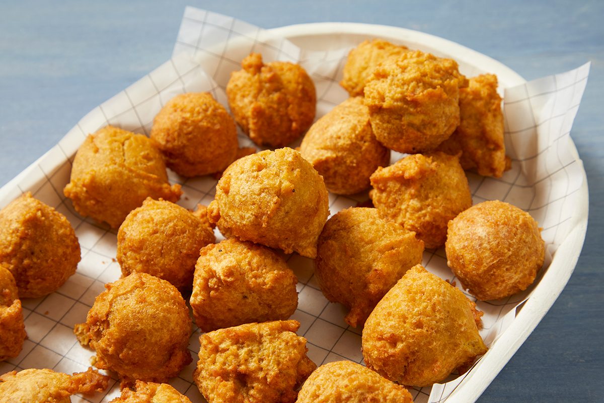 A tray filled with golden-brown, crispy hush puppies arranged neatly on a checkered paper. The hush puppies appear freshly fried, exhibiting a textured surface against a light blue background.