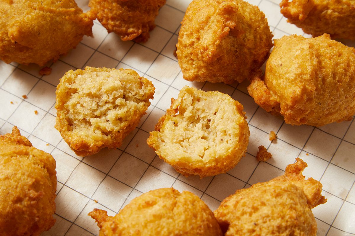 Golden brown hush puppies on a white grid-patterned surface. Some hush puppies are whole while others are cut in half, revealing their soft, moist interior. The texture looks crispy on the outside.