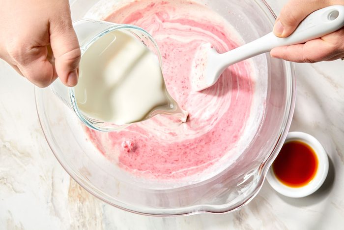 A person pouring whipping cream into a bowl of raspberries and sugar.