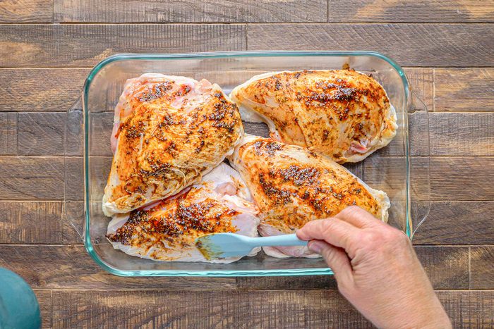 overhead shot of a glass baking dish with four raw chicken breasts inside; the chicken breasts are coated in a brown marinade; a hand holds a blue silicone spatula and spreads the marinade onto the chicken breasts; the baking dish is sitting on a wooden surface
