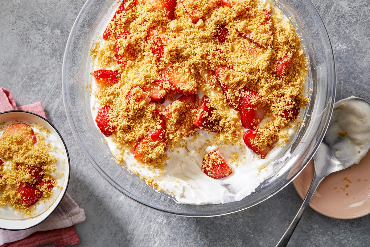 A large bowl of strawberry shortcake dip with crumbled topping and sliced strawberries on a creamy base. A smaller bowl with a serving of the dip is nearby on a gray surface with a pink plate and spoon.