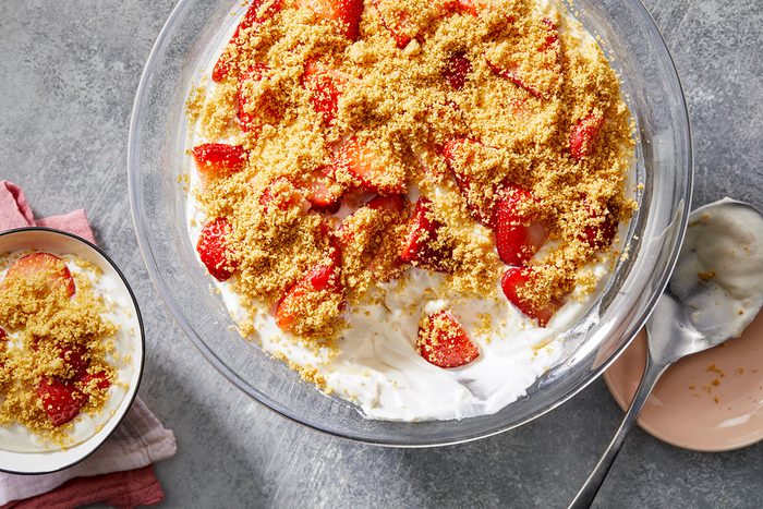 A large bowl of strawberry shortcake dip with crumbled topping and sliced strawberries on a creamy base. A smaller bowl with a serving of the dip is nearby on a gray surface with a pink plate and spoon.