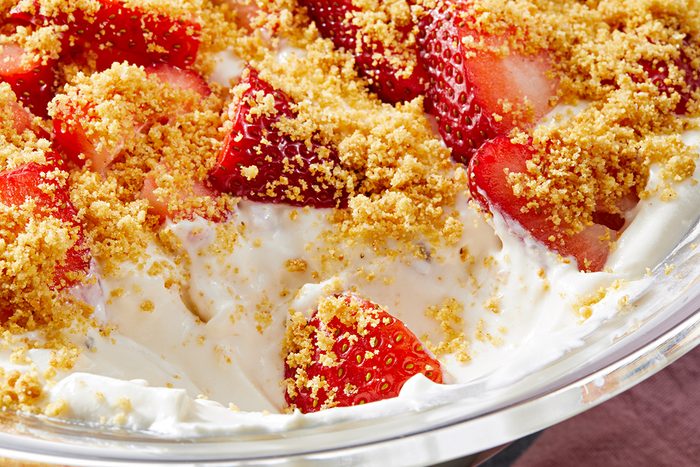 A close-up of a dessert featuring fresh strawberry slices layered with creamy white filling, topped with golden-brown crumble in a glass dish. A spoonful appears to have been taken from the center.
