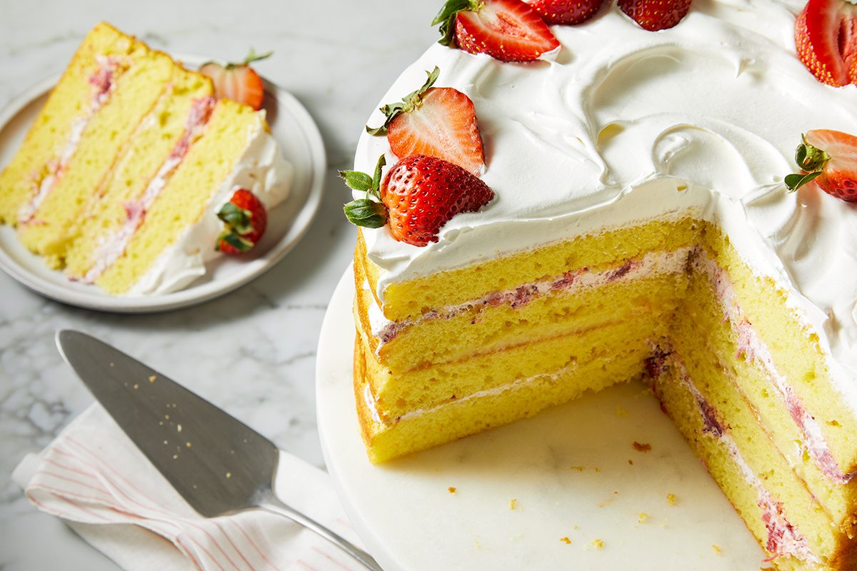 A sliced layer cake with yellow sponge and pink filling, topped with white frosting and fresh strawberry halves. A slice sits on a separate plate beside a cake server and napkin, all set on a marble surface.