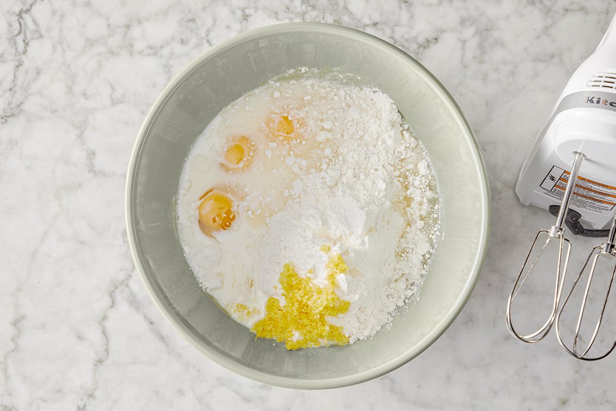 A green bowl on a marble countertop contains ingredients for baking: cracked eggs, flour, sugar, butter, and milk. A white electric mixer with metal beaters is positioned next to the bowl.