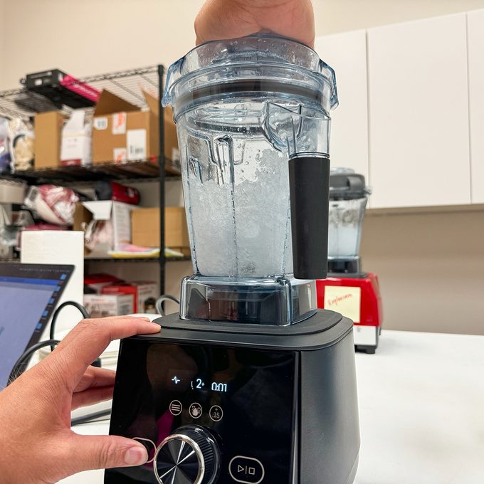 A person demonstrates blending in a transparent blender pitcher, filled with water, on a black base. Shelving and a red appliance are visible in the background.