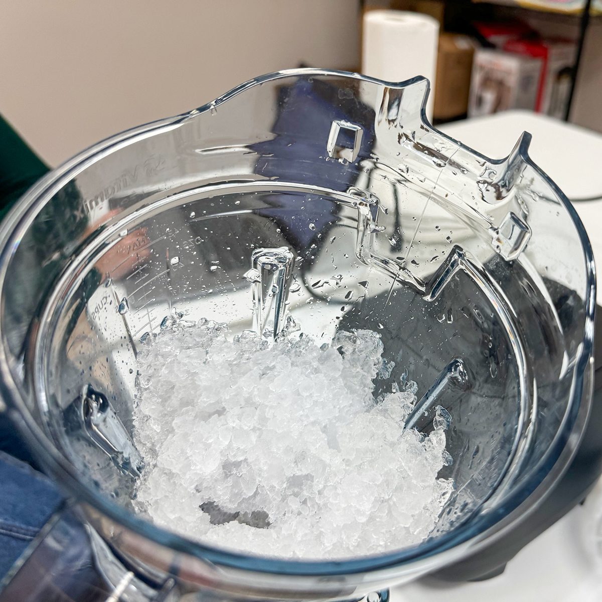 A close-up view of crushed ice inside a transparent blender jug. The jug is placed on a countertop, with a glimpse of a green sleeve on the left. The background includes some shelves with various items.
