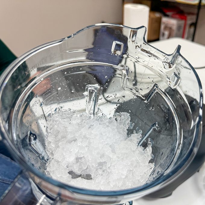 A close-up view of crushed ice inside a transparent blender jug. The jug is placed on a countertop, with a glimpse of a green sleeve on the left. The background includes some shelves with various items.