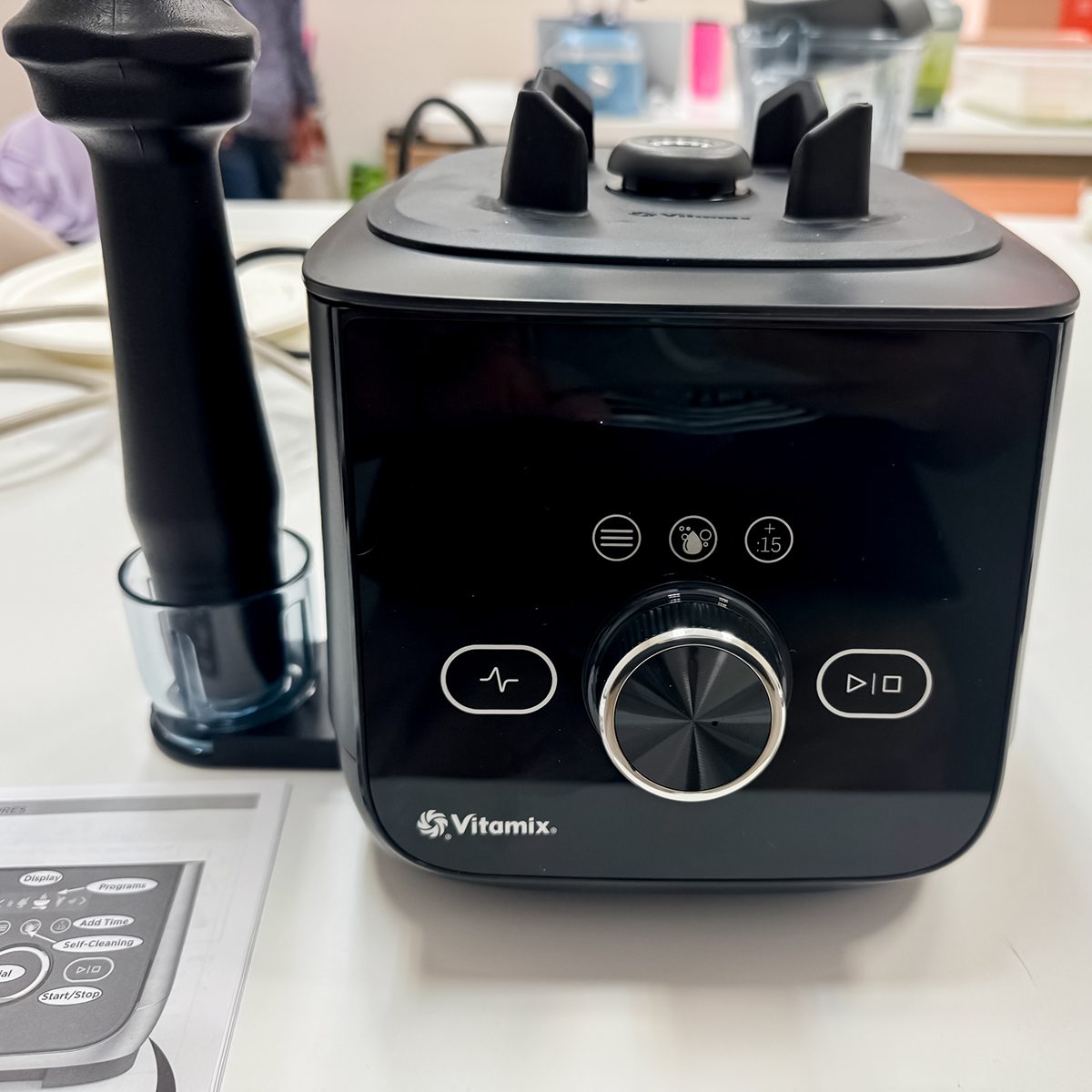 A black Vitamix blender with control knobs and buttons is placed on a white surface. An attachment is secured on the left side of the base. A blurred background and a paper with a printed image of the blender are visible.