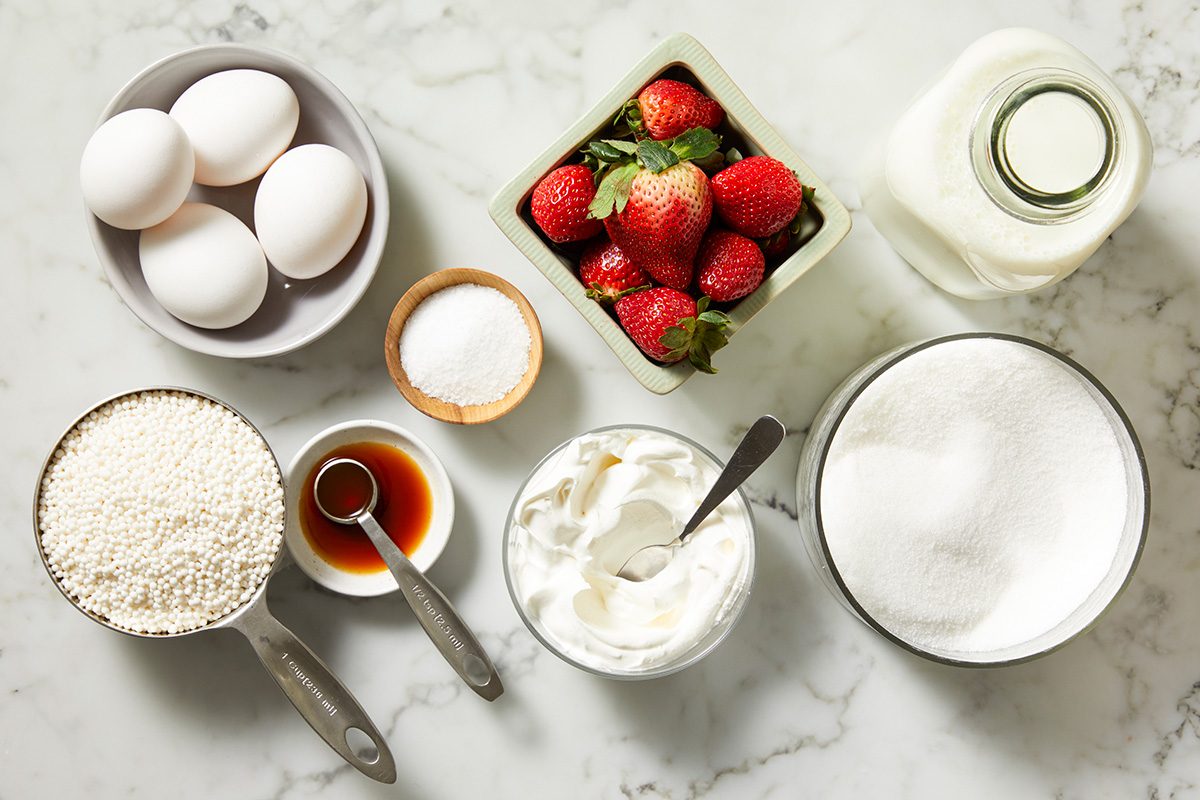 Baking ingredients on a marble countertop include eggs, strawberries, a bottle of milk, sugar in a bowl, a measuring cup of flour, a bowl of whipped cream, a small bowl of vanilla extract, and a bowl of salt.