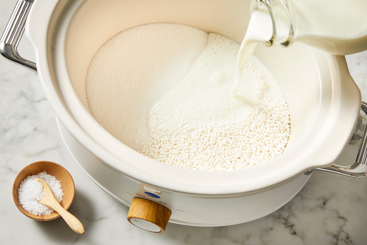 A white crockpot on a marble surface containing sugar is being filled with milk from a glass bottle. A small wooden bowl with salt and a spoon is beside the crockpot.