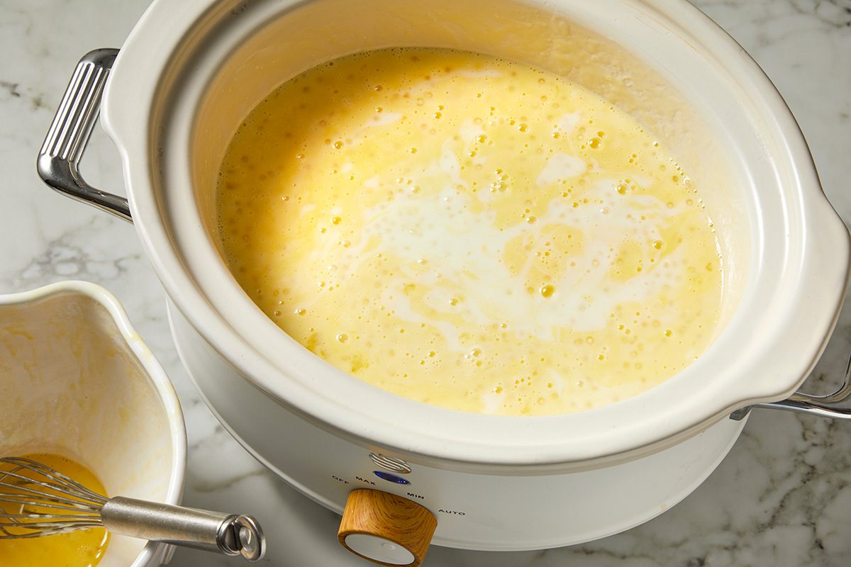 A slow cooker filled with a creamy mixture sits on a marble countertop. Next to it, a small bowl with a whisk rests, suggesting preparation of a recipe. The mixture appears to be light yellow and frothy.