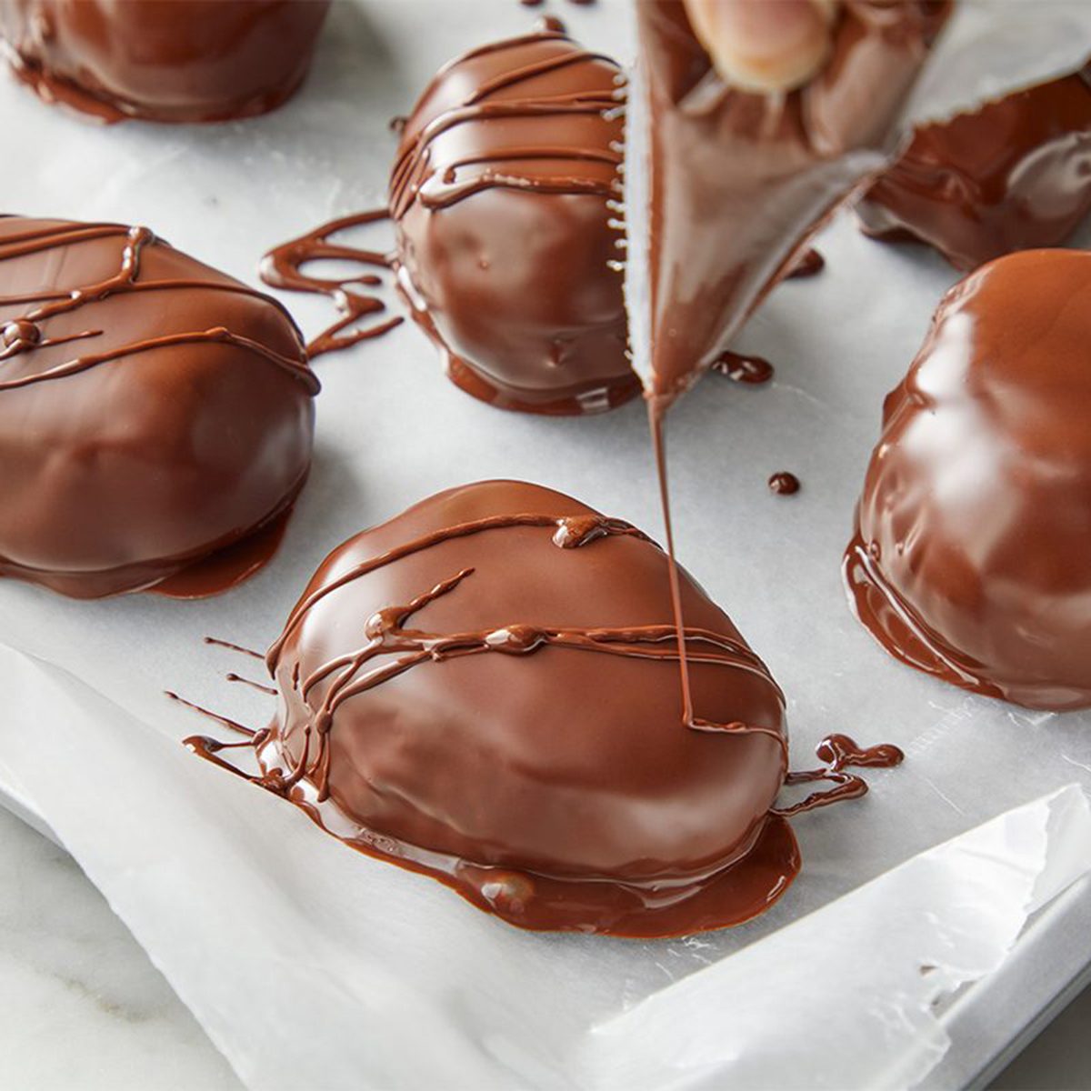 A person drizzles chocolate over several chocolate-covered treats on parchment paper. The treats are oval-shaped, glossy, and placed on a baking tray.