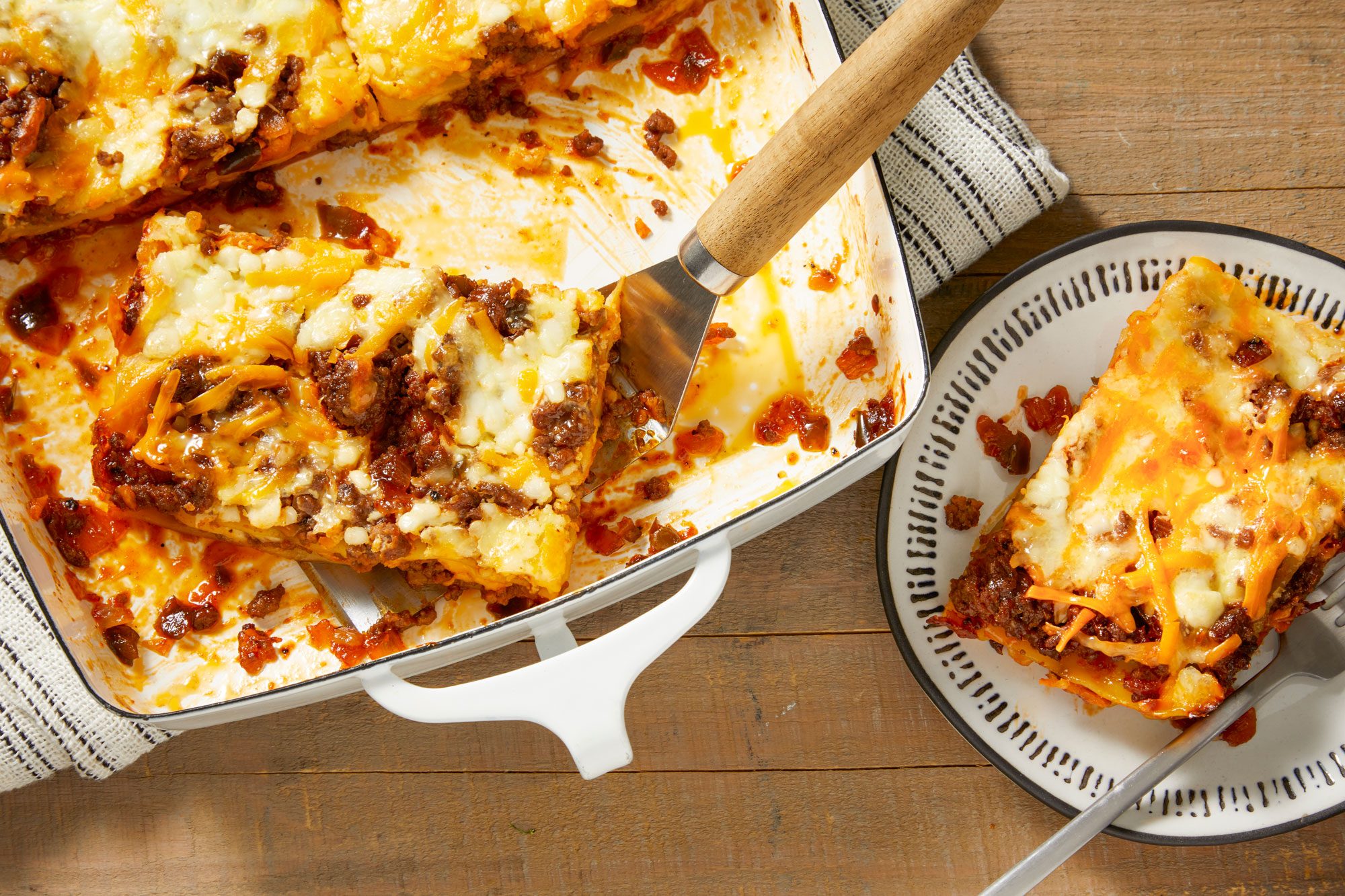 overhead shot of Barbecue Lasagna, which is presented in a rectangular baking dish; The lasagna is partially cut, A serving has been placed on a circular plate beside the dish, A spatula rests in the baking dish, while a striped kitchen towel can be seen under the dish on the wooden surface