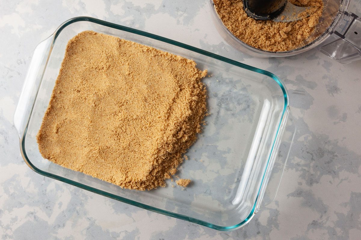overhead shot of a glass baking dish filled with a crushed graham crackers or a similar ingredient; to the side, a food processor can be seen, containing more of the same crumbly mixture, the background features a light, textured surface