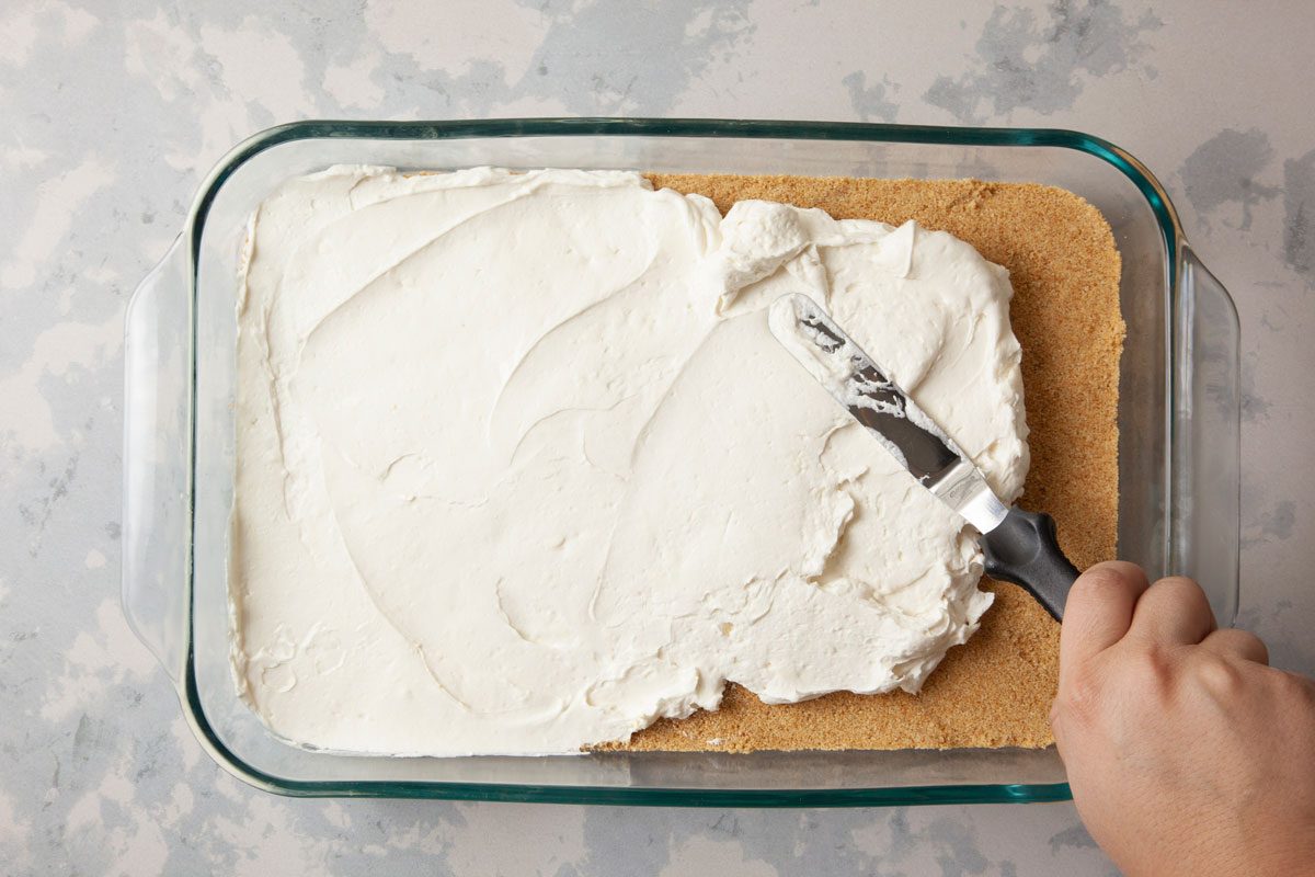 overhead shot of a rectangular glass baking dish containing a layer of white cream topping spread over a crumbly base; a hand holding a spatula is seen in the process of smoothing out the cream; the background is a light gray color