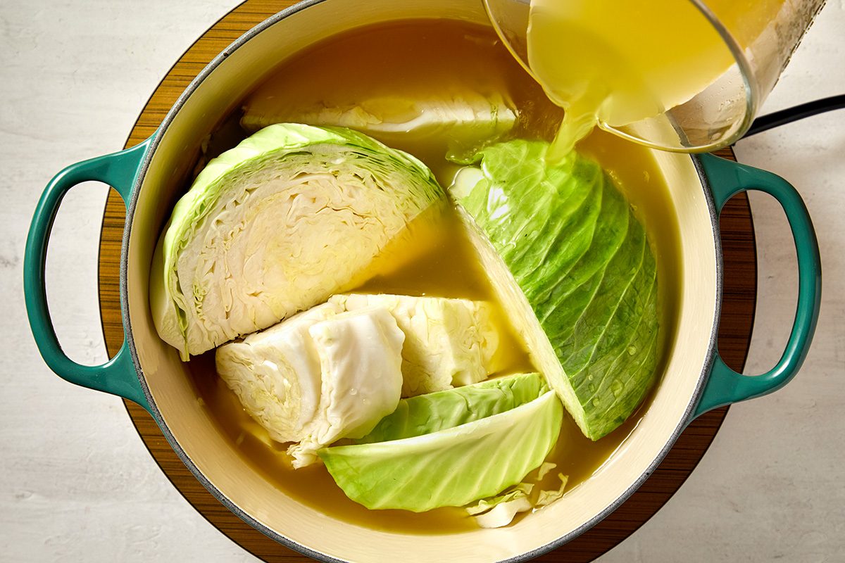 A pot filled with cabbage leaves in broth, with more broth being poured in from a container. The pot has teal handles and is placed on a wooden trivet against a light background.