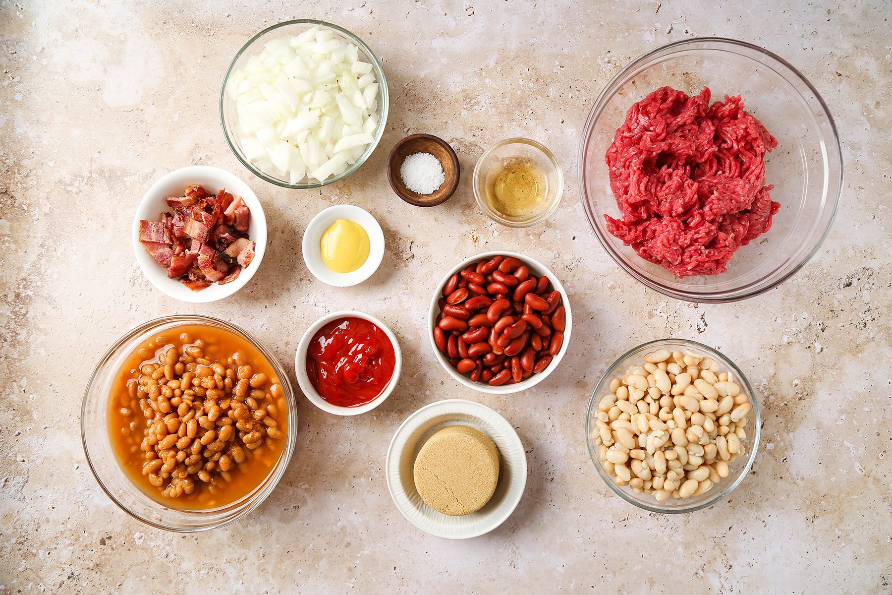 Ingredients for a chili recipe arranged on a countertop include chopped onions, ground beef, bacon bits, white beans, kidney beans, baked beans, mustard, ketchup, brown sugar, and seasonings.