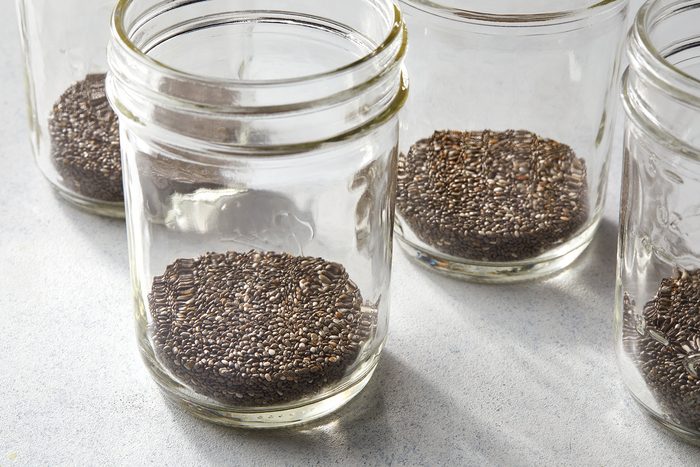 Four clear glass jars each containing a portion of black chia seeds placed on a light gray surface. The jars are open, with the chia seeds filling the bottom of each jar.