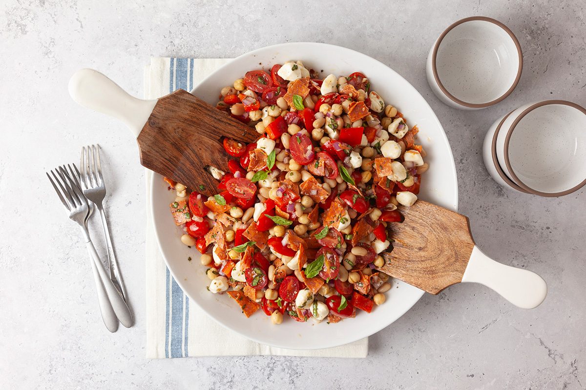 A bowl of colorful salad with cherry tomatoes, chickpeas, mozzarella balls, and herbs, served on a white plate with two wooden-handled serving utensils, next to stacked plates and forks on a blue-striped napkin.