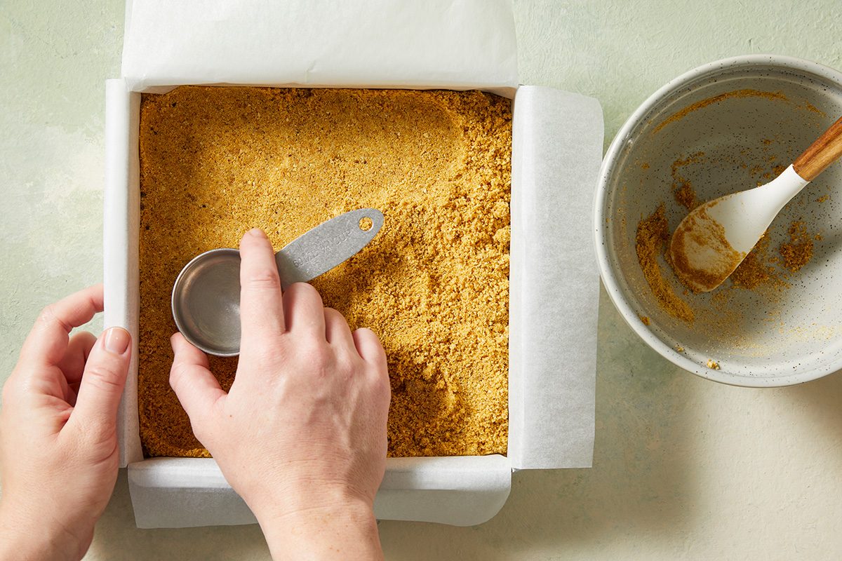 A person presses a crumbly mixture into a parchment-lined square baking pan using a metal measuring cup. A mixing bowl with a spatula and remaining crumbs sits nearby on a light-colored surface.