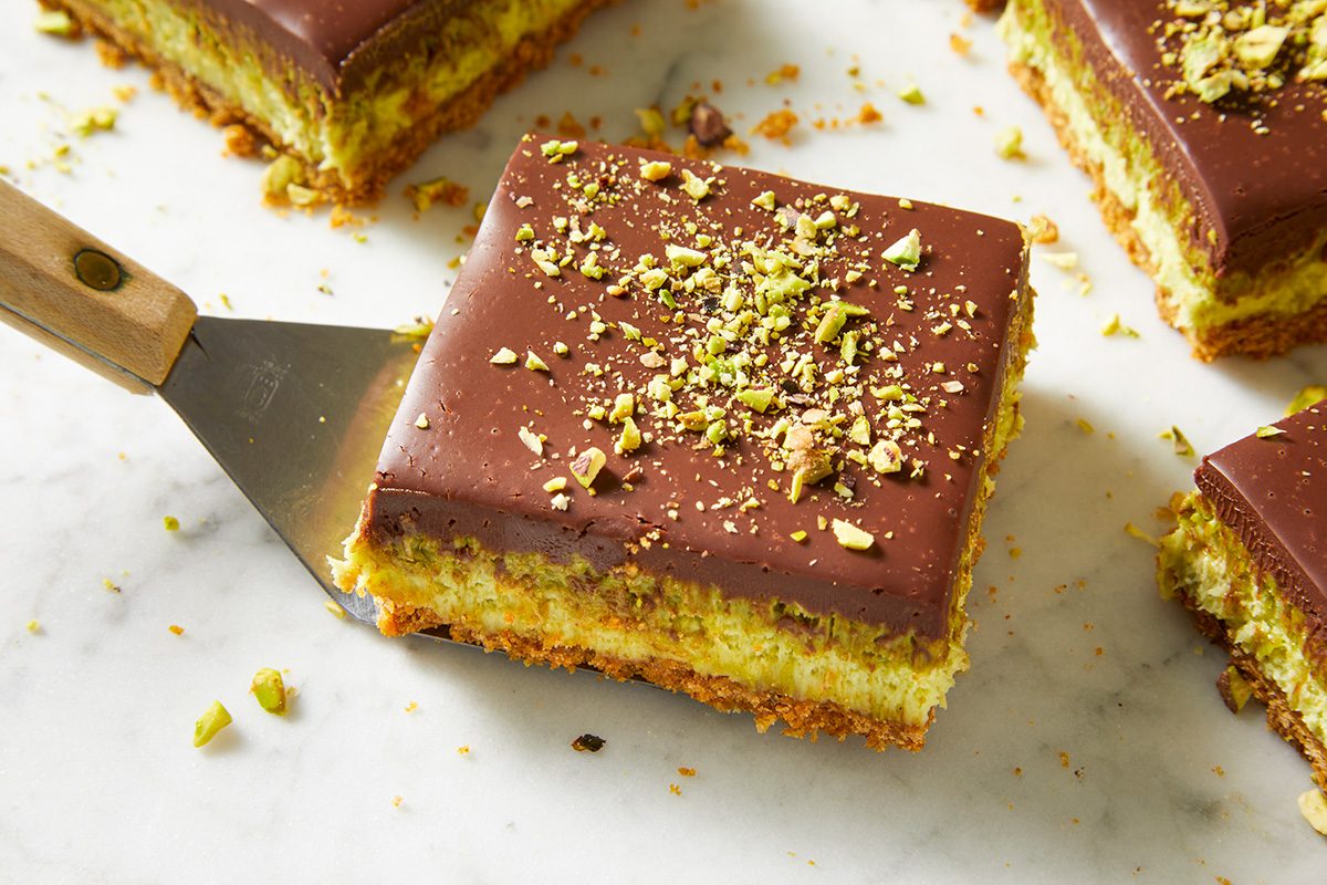 A spatula lifts a square dessert bar with a chocolate topping, chopped pistachios, and layered cookie and creamy filling on a marble surface. Other dessert bars are visible in the background.